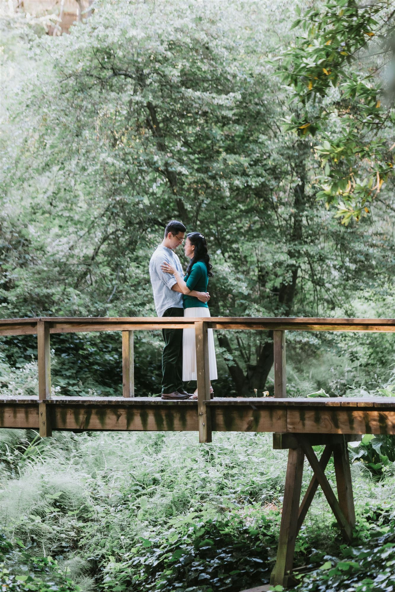 Helen and Richard embracing on a wooden footbridge surrounded by ferns and trees